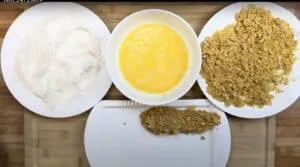 Breading process with flour, beaten eggs, and cornflakes for crispy coating on a wooden cutting board.